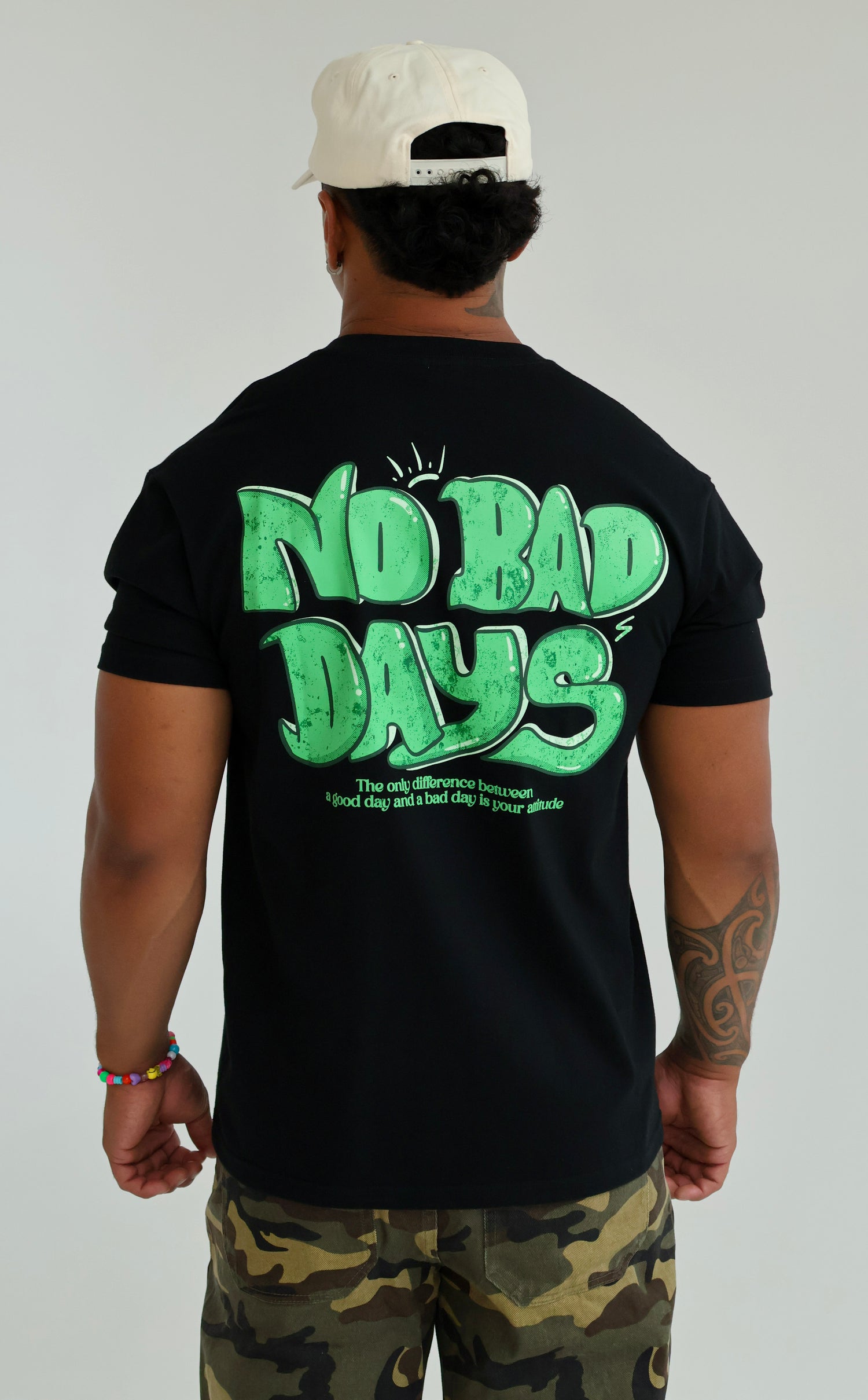 Man wearing black graphic t-shirt with green 'No Bad Days' text, camo pants, and white cap
