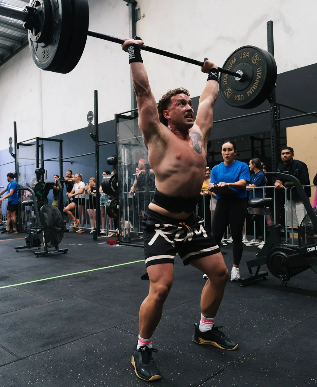 Man lifting heavy barbell overhead during intense CrossFit workout in gym with spectators