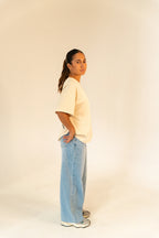 Woman in beige oversized t-shirt, light blue wide-leg jeans, and sneakers on plain background