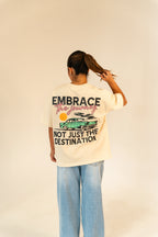 Woman with ponytail wearing a beige t-shirt with car and text Embrace the journey, not just the destination