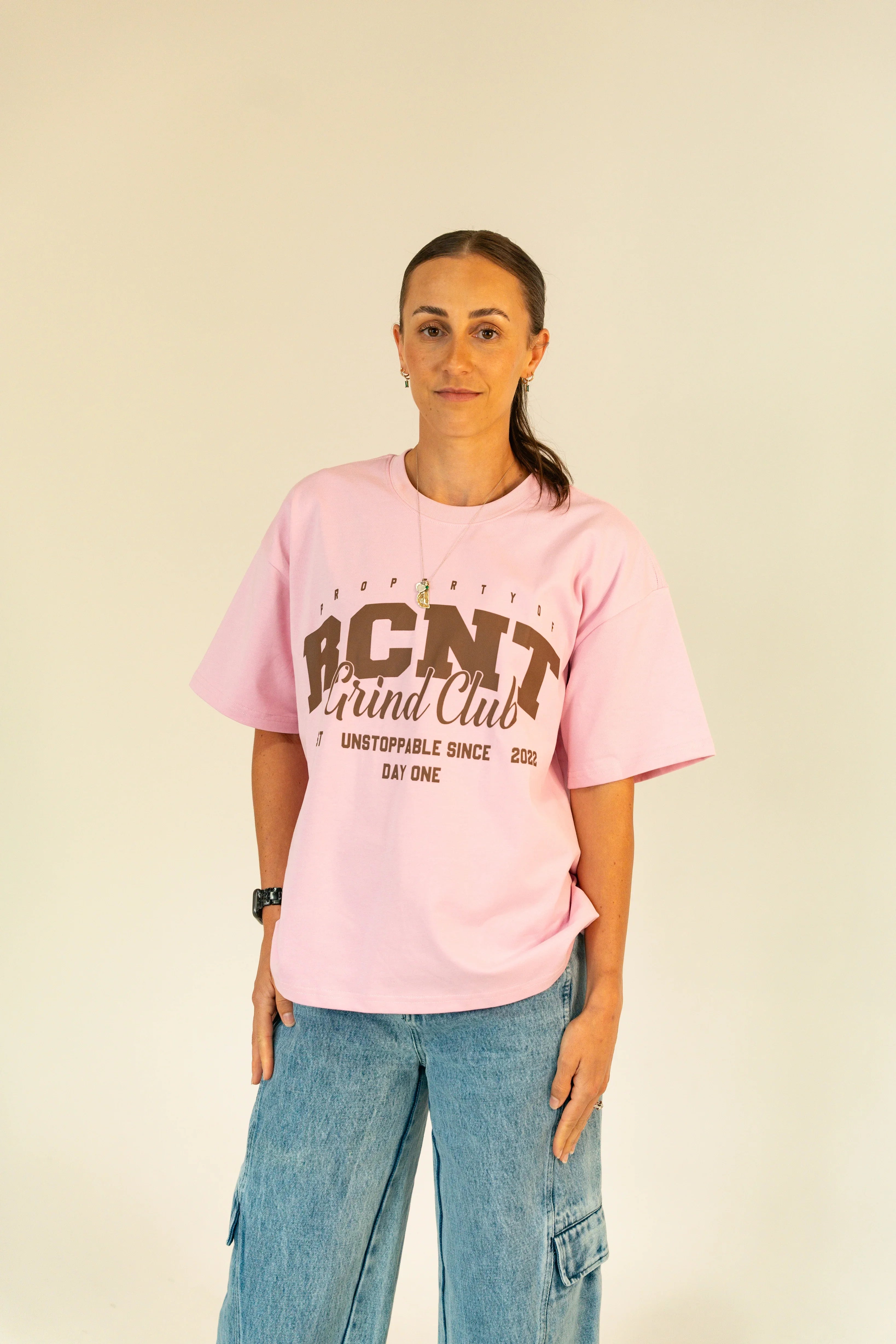 Woman wearing pink RCNT Grind Club t-shirt and blue cargo jeans against light background