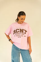 Woman wearing a pink RCNT Grind Club graphic t-shirt and blue denim jeans against beige background