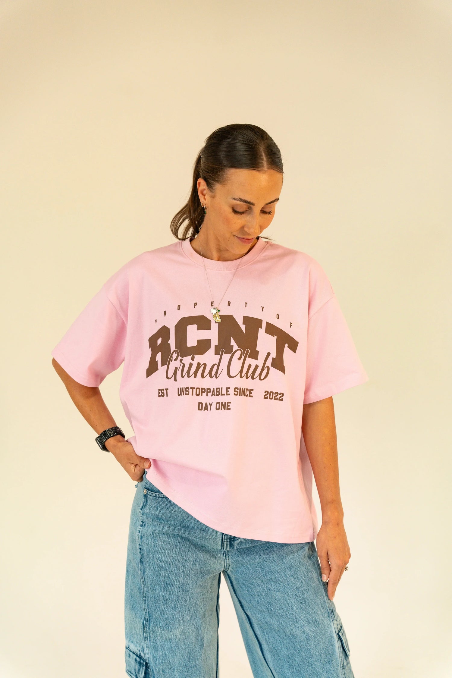 Woman wearing a pink RCNT Grind Club graphic t-shirt and blue denim jeans against beige background