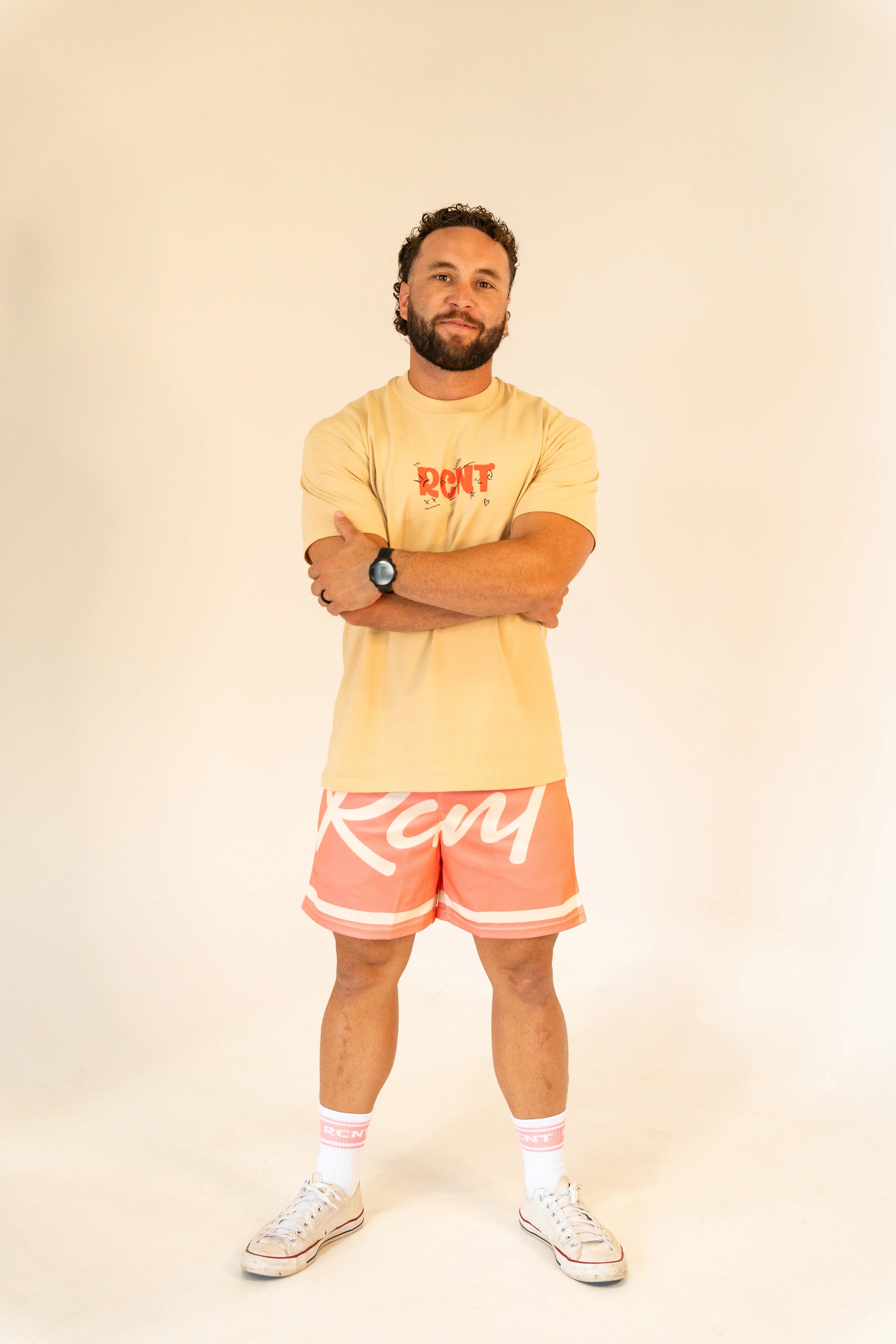 Man in casual beige RCN t-shirt, coral RCN shorts, white RCN socks, and white sneakers standing with arms crossed