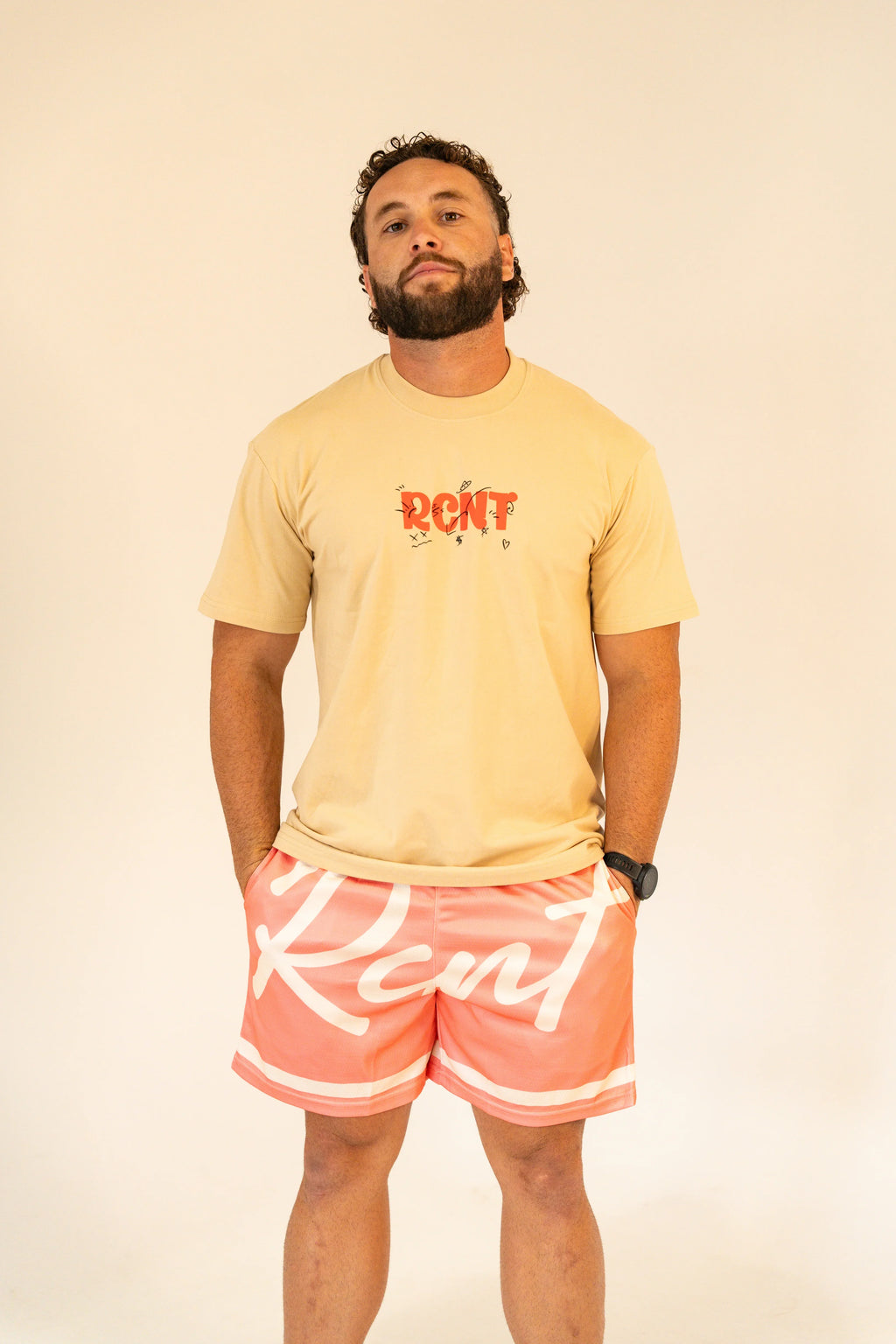 Man wearing beige graphic t-shirt and coral shorts with white script lettering against plain background