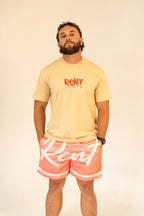 Man wearing beige graphic t-shirt and coral shorts with white script lettering against plain background