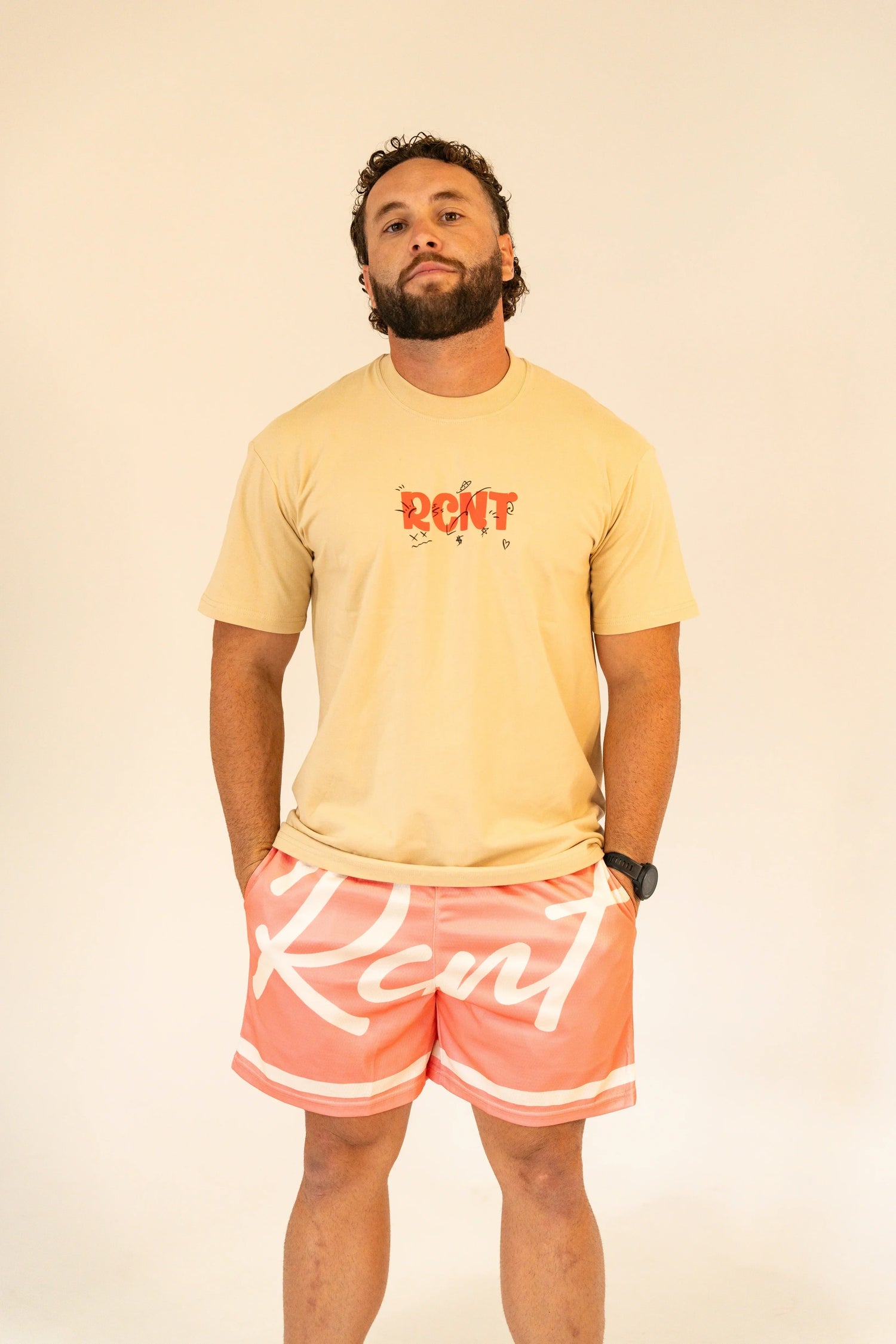 Man wearing beige graphic t-shirt and coral shorts with white script lettering against plain background