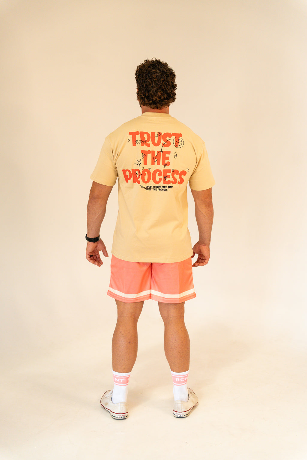 Man wearing beige T-shirt with 'Trust The Process' text and coral shorts in studio