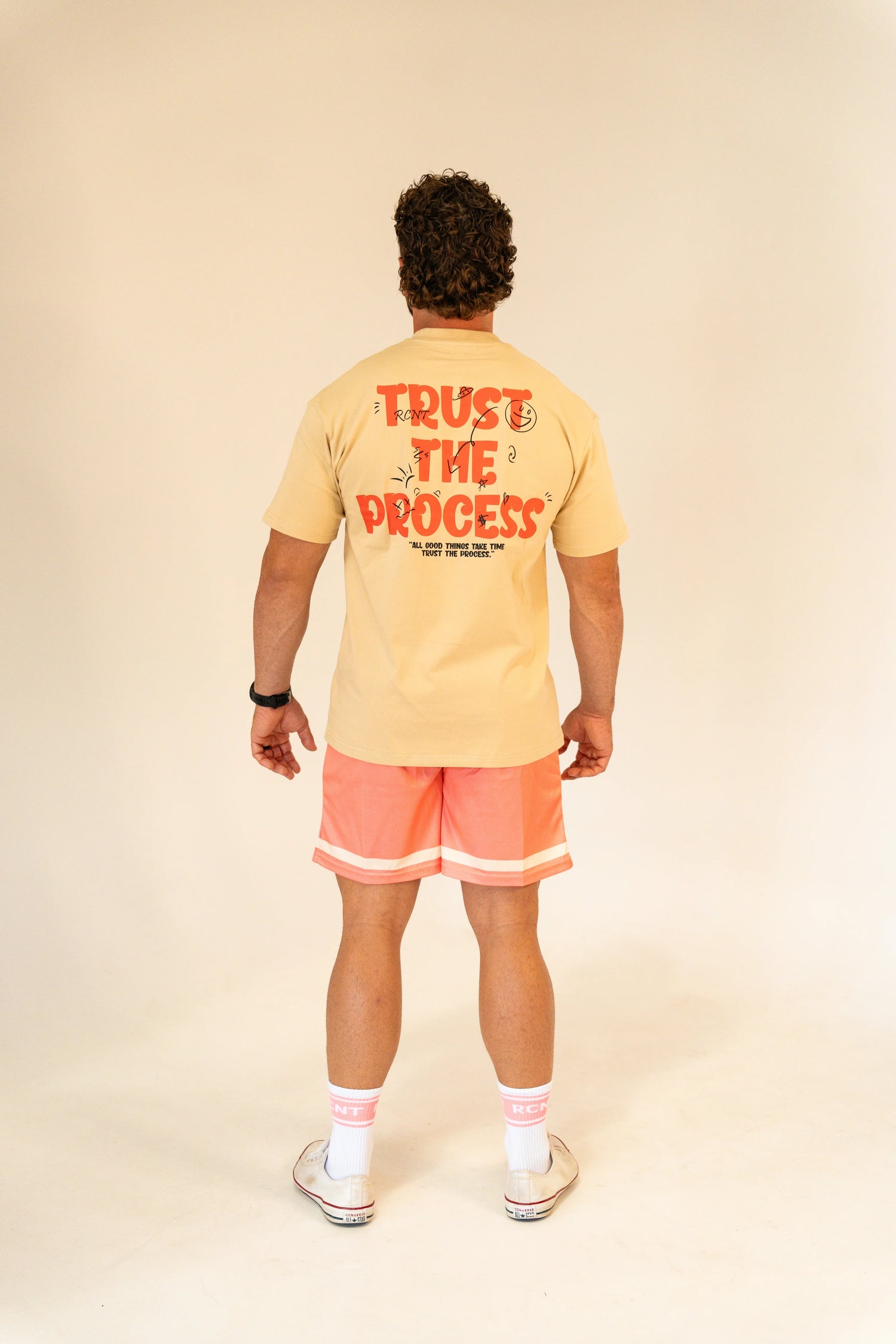 Man wearing beige T-shirt with 'Trust The Process' text and coral shorts in studio