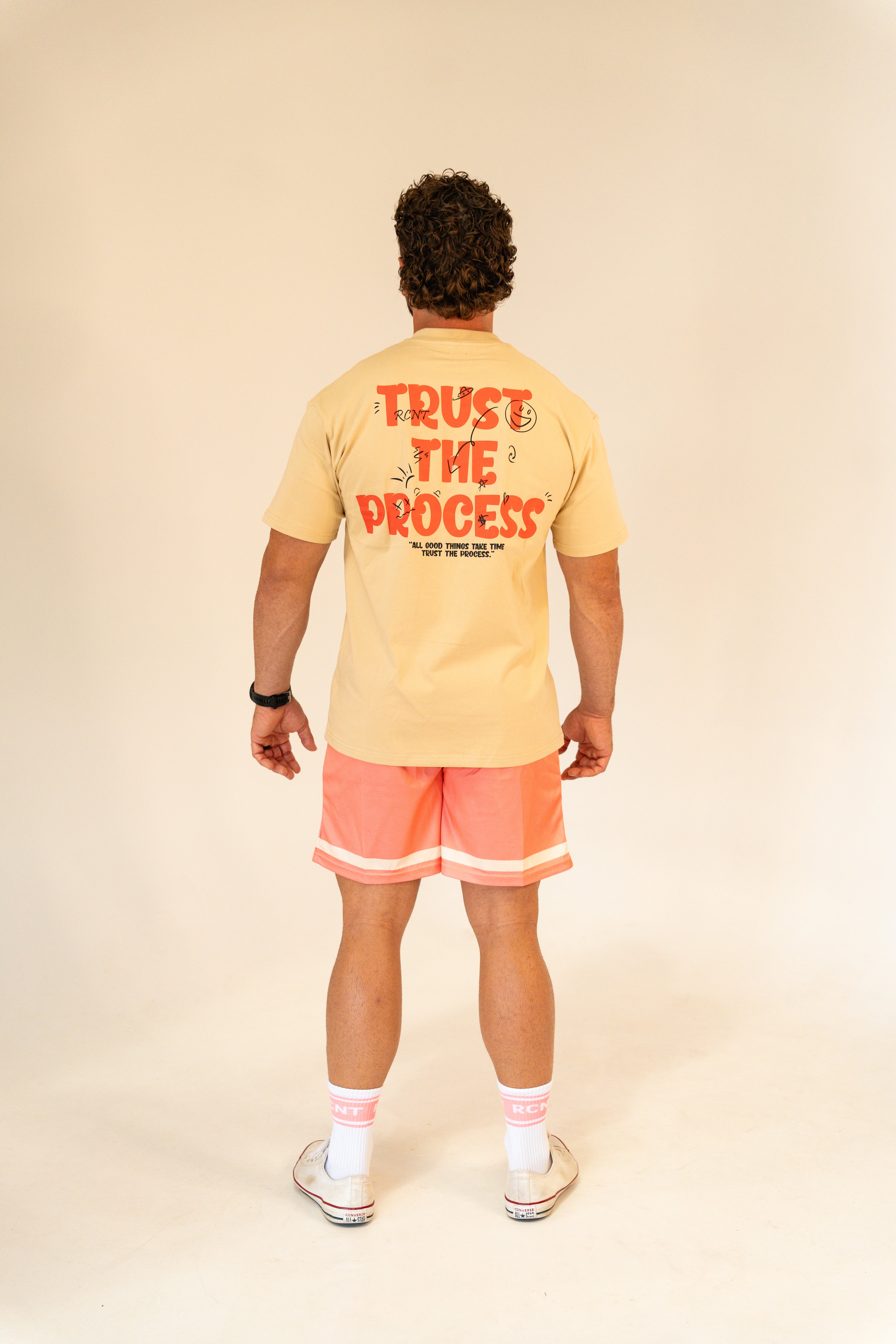 Man wearing beige T-shirt with 'Trust The Process' text and coral shorts in studio
