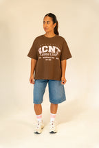 Woman wearing brown BCNT Grind Club graphic t-shirt with denim shorts and white sneakers