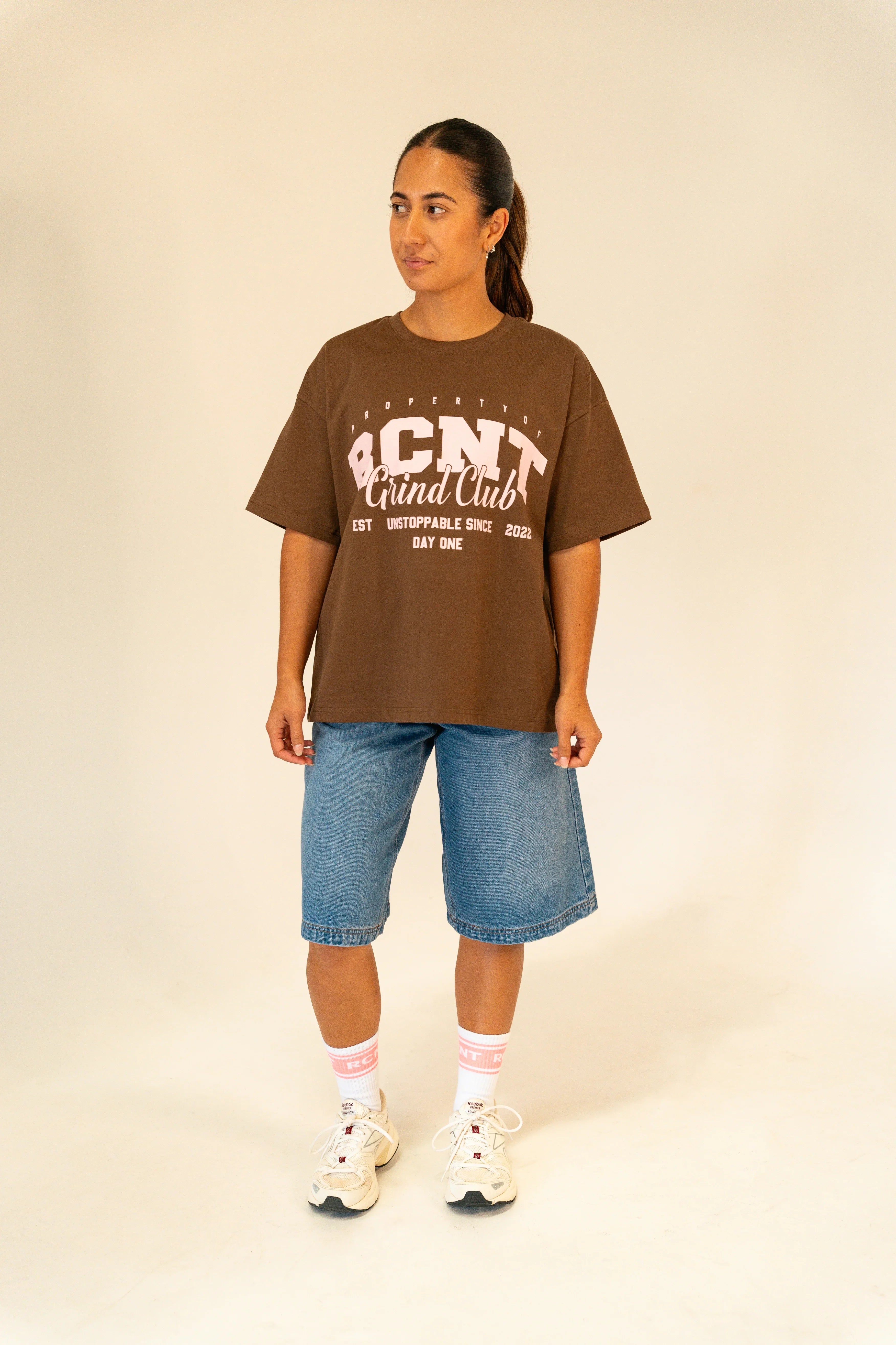 Woman wearing brown BCNT Grind Club graphic t-shirt with denim shorts and white sneakers