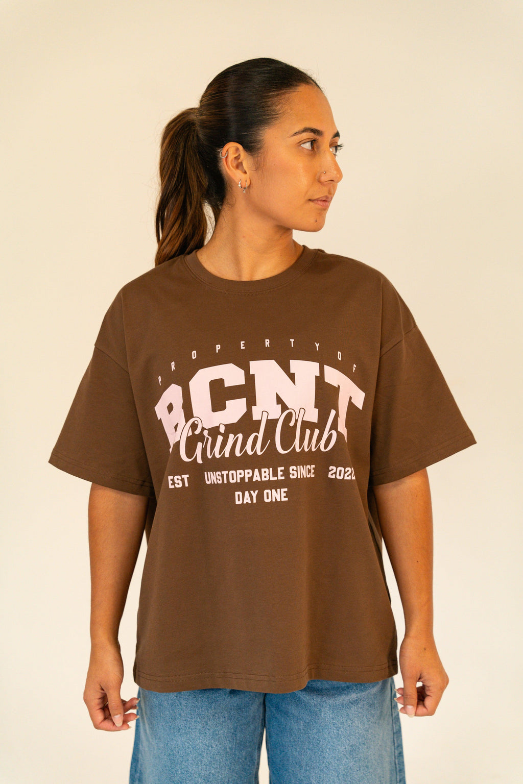 Woman wearing brown BCNT Grind Club graphic t-shirt and blue jeans standing against a plain background