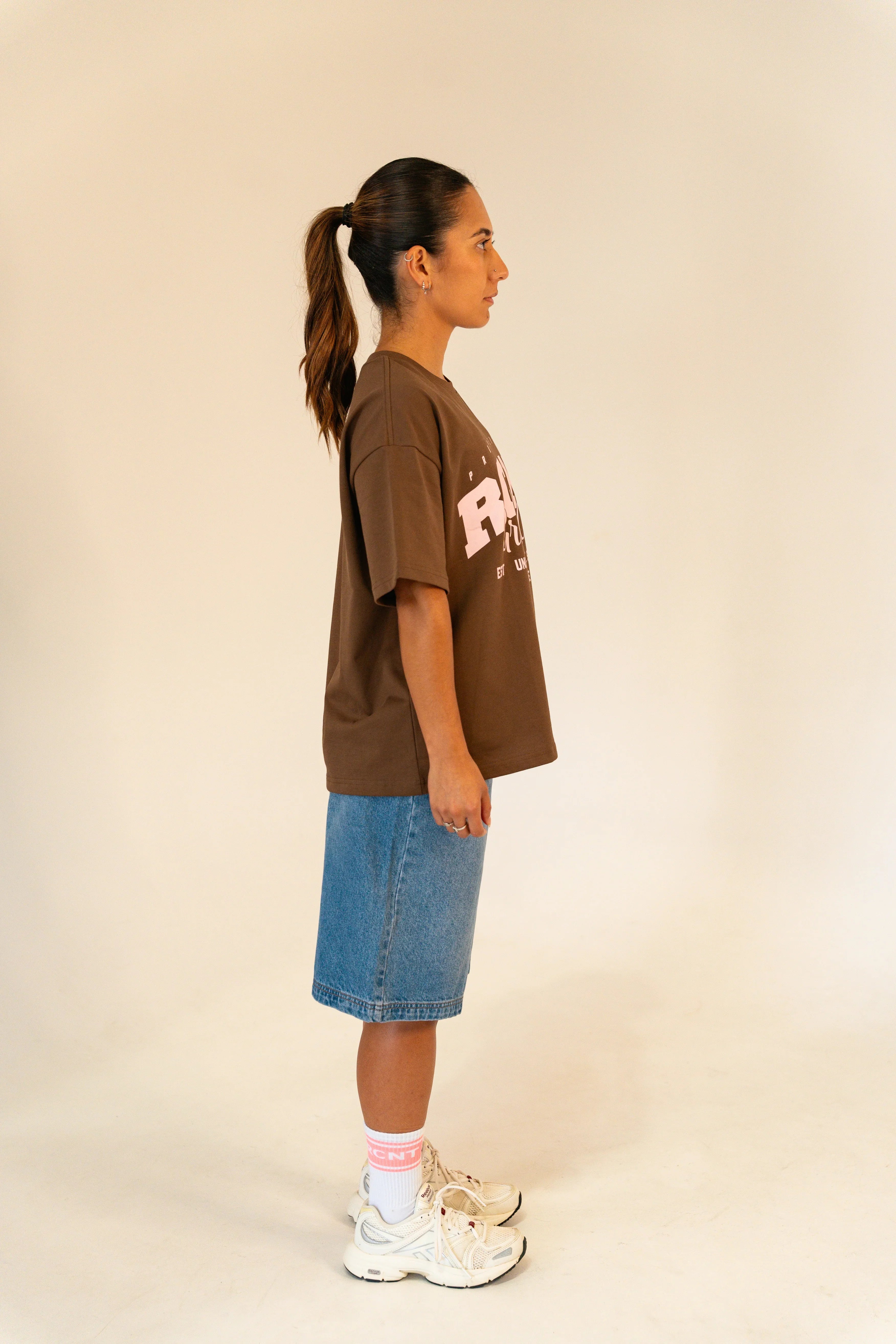 Woman in brown graphic t-shirt, denim skirt, pink socks, and white sneakers standing sideways on beige background