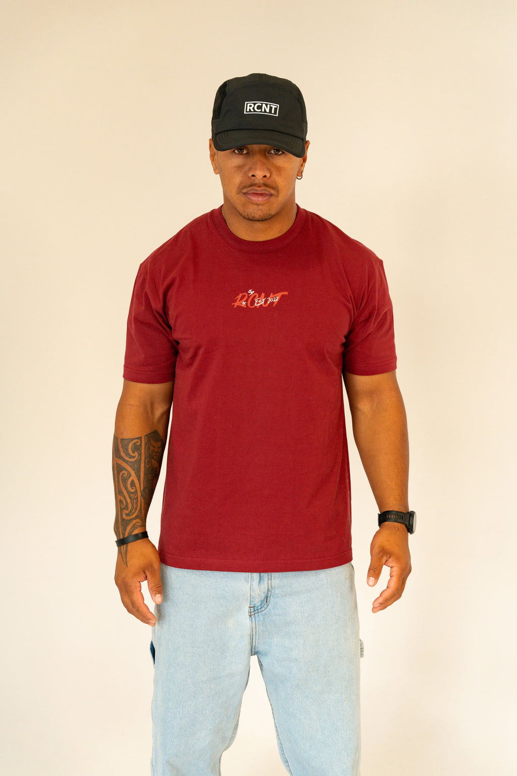 Man wearing maroon RCNT T-shirt and black RCNT cap with light blue jeans on beige background