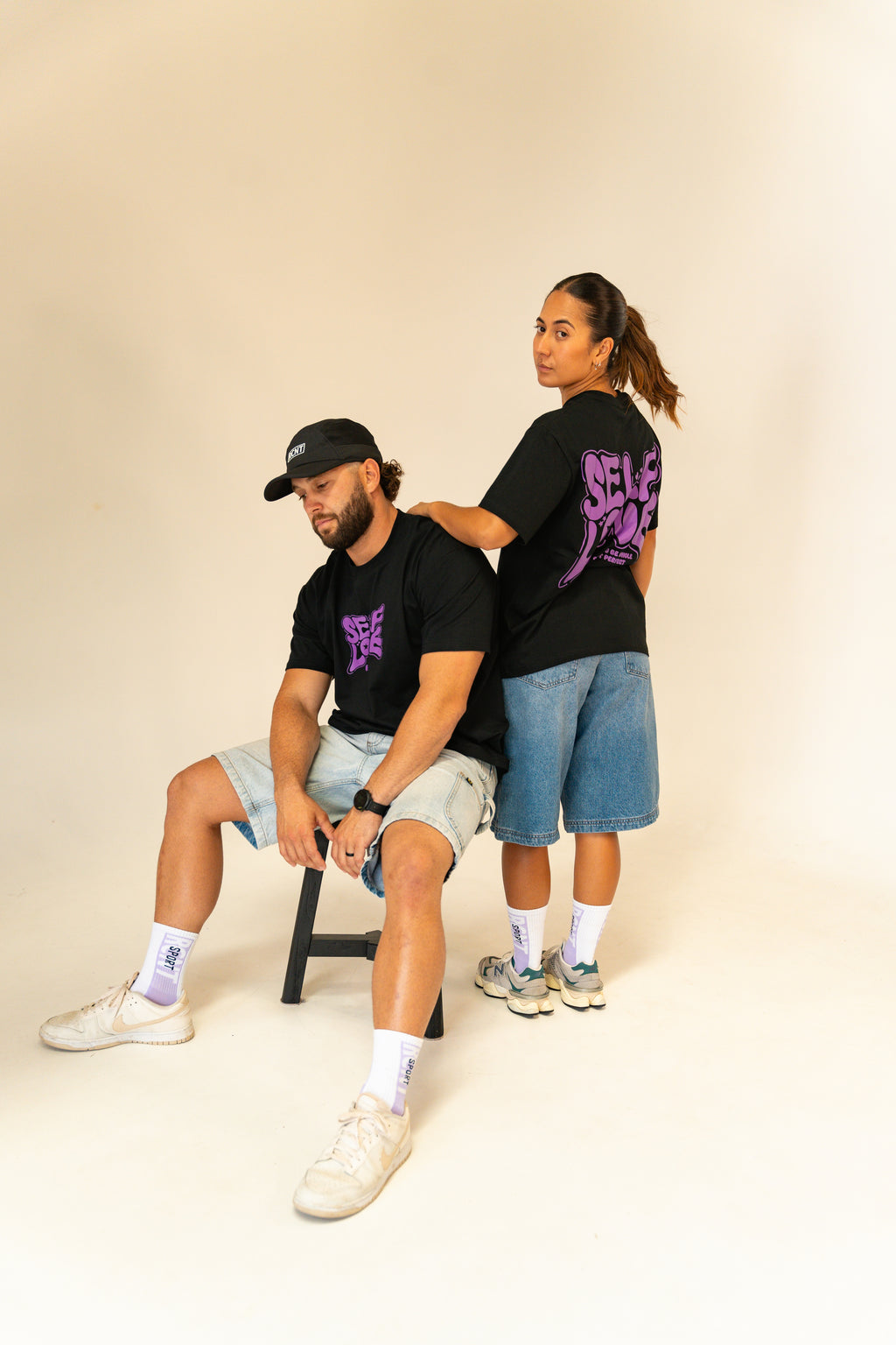 Man sitting and woman standing in black graphic t-shirts with purple text, denim shorts, and casual sneakers on white background