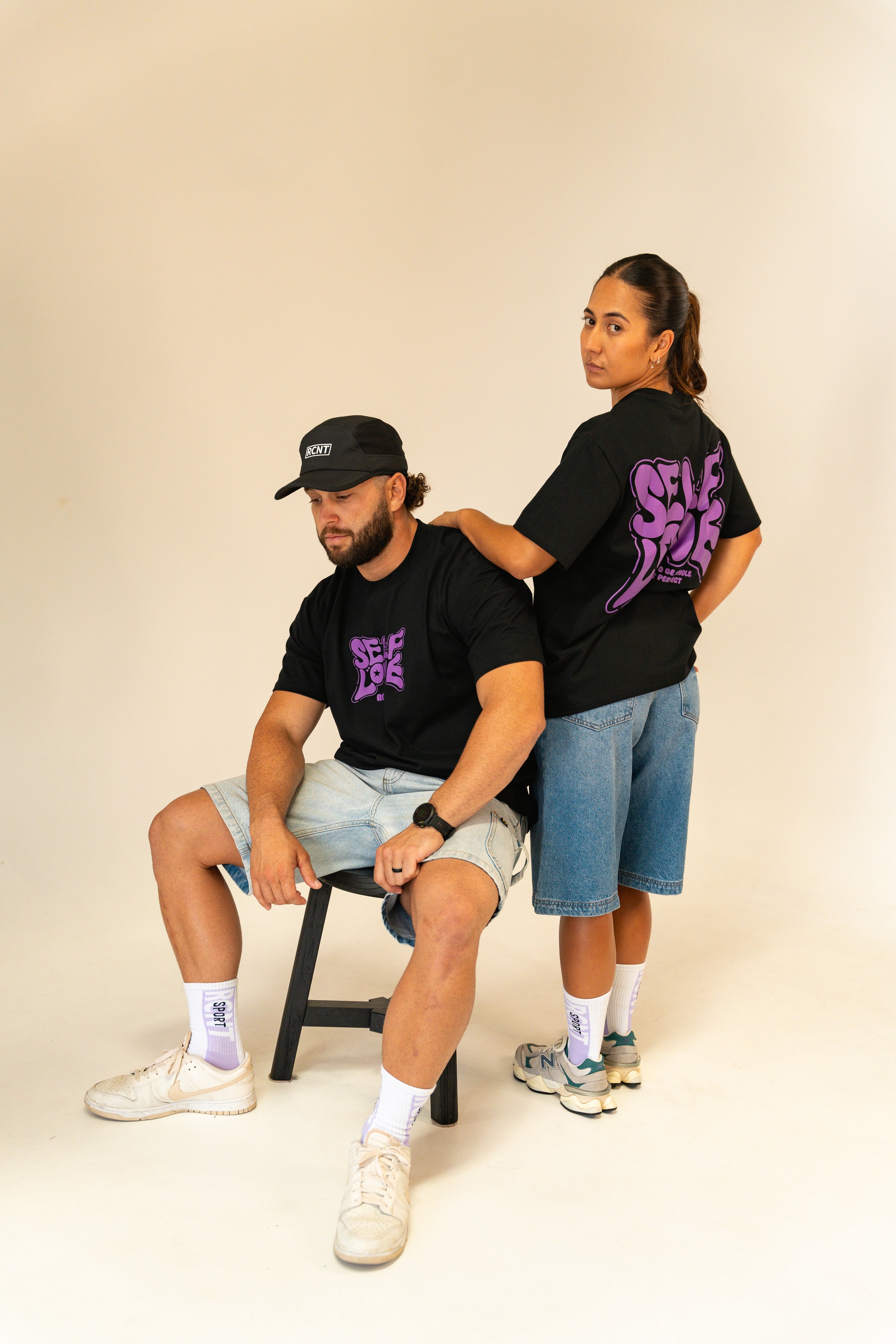 Man seated and woman standing wearing black self love graphic t-shirts and denim shorts in studio
