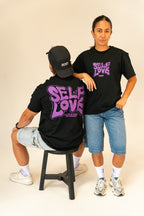 Man sitting on stool and woman standing wearing black Self Love graphic t-shirts and denim shorts in studio
