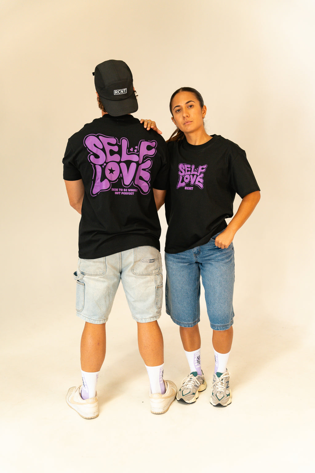 Two models wearing black self love graphic t-shirts with purple text, denim shorts, and white sneakers in studio