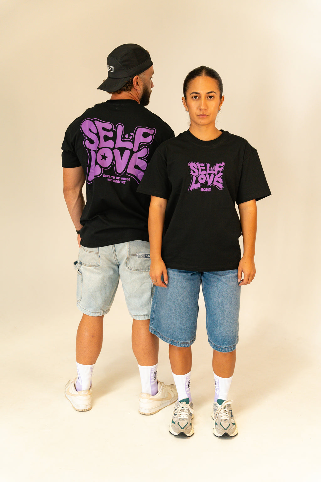 Man and woman modeling black t-shirts with purple "Self Love" graphic, paired with denim shorts in studio