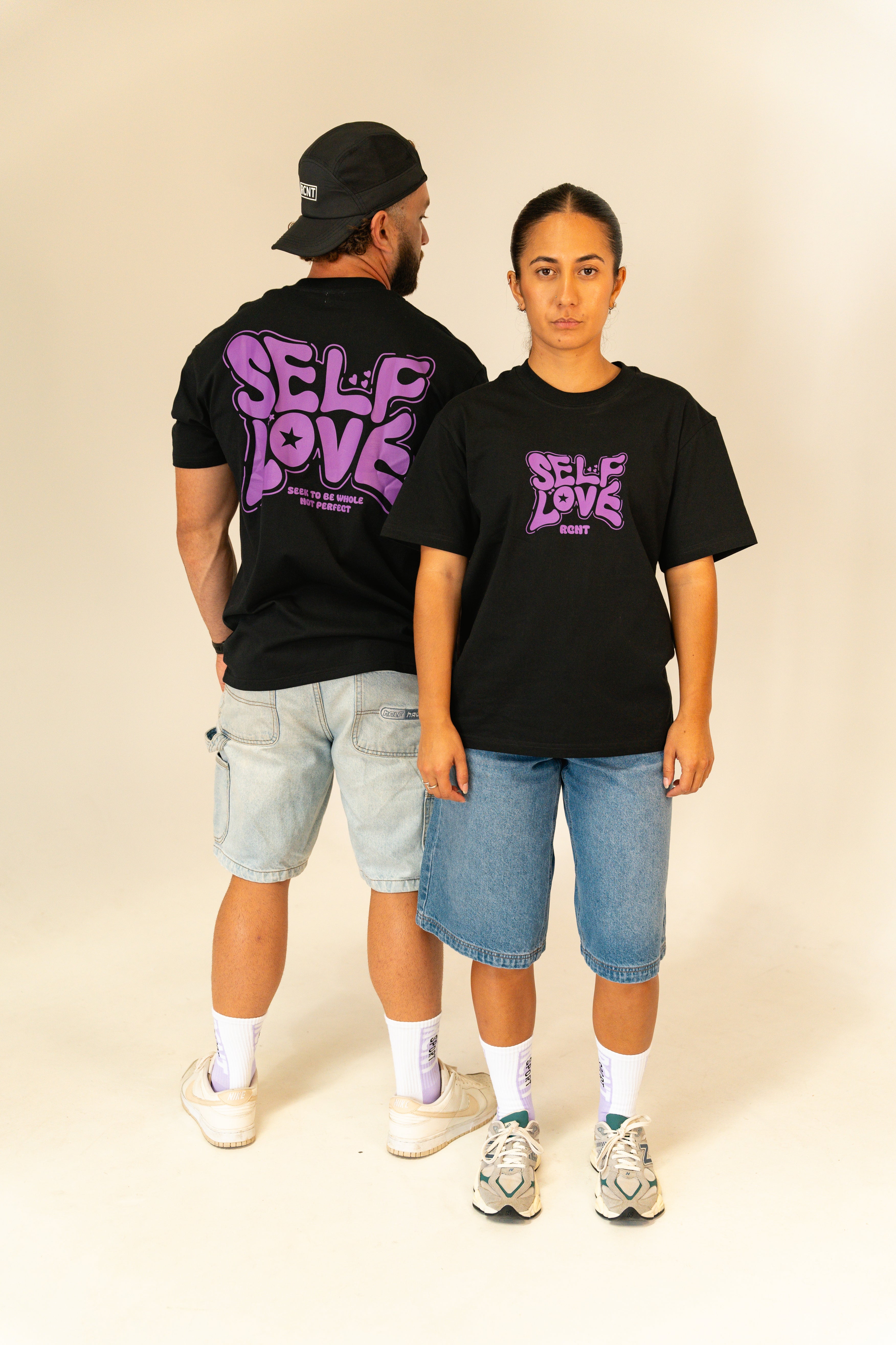 Man and woman modeling black t-shirts with purple "Self Love" graphic, paired with denim shorts in studio