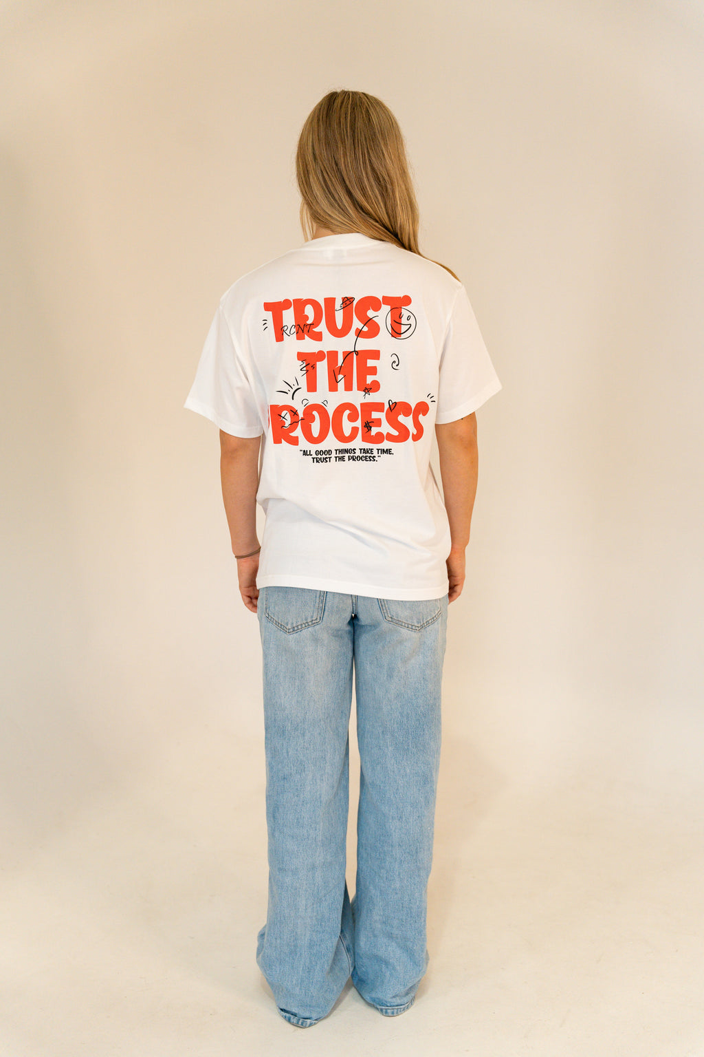 Person wearing white T-shirt with red text 'Trust the Process' and blue jeans against plain background