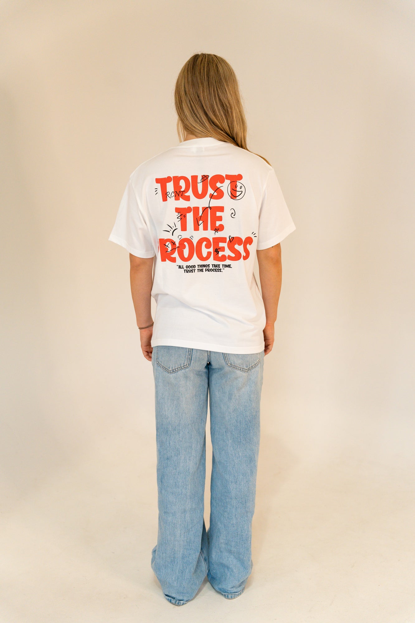 Person wearing white T-shirt with red text 'Trust the Process' and blue jeans against plain background