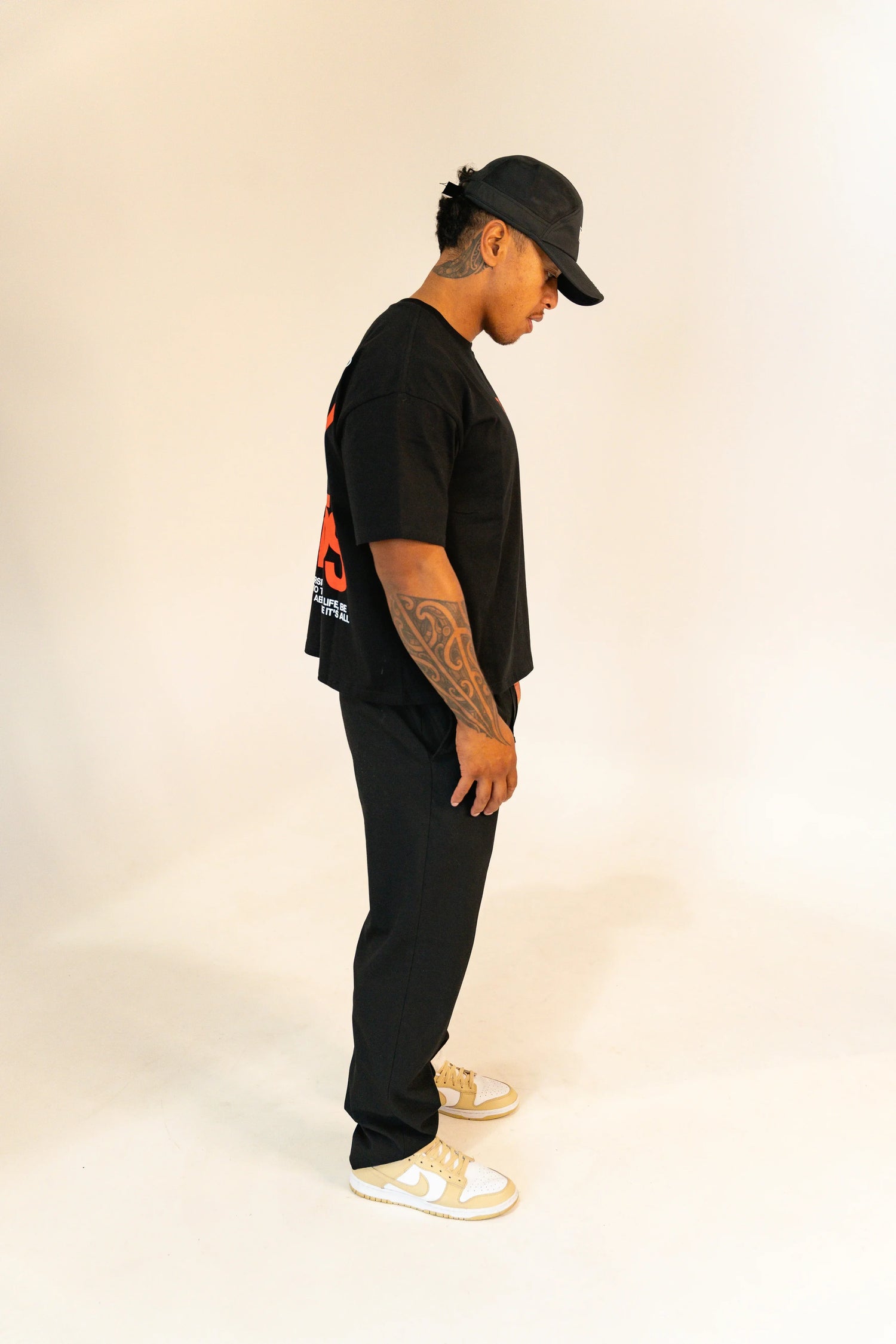 Man in black cap, black graphic t-shirt, black pants, and beige and white Nike sneakers on white background