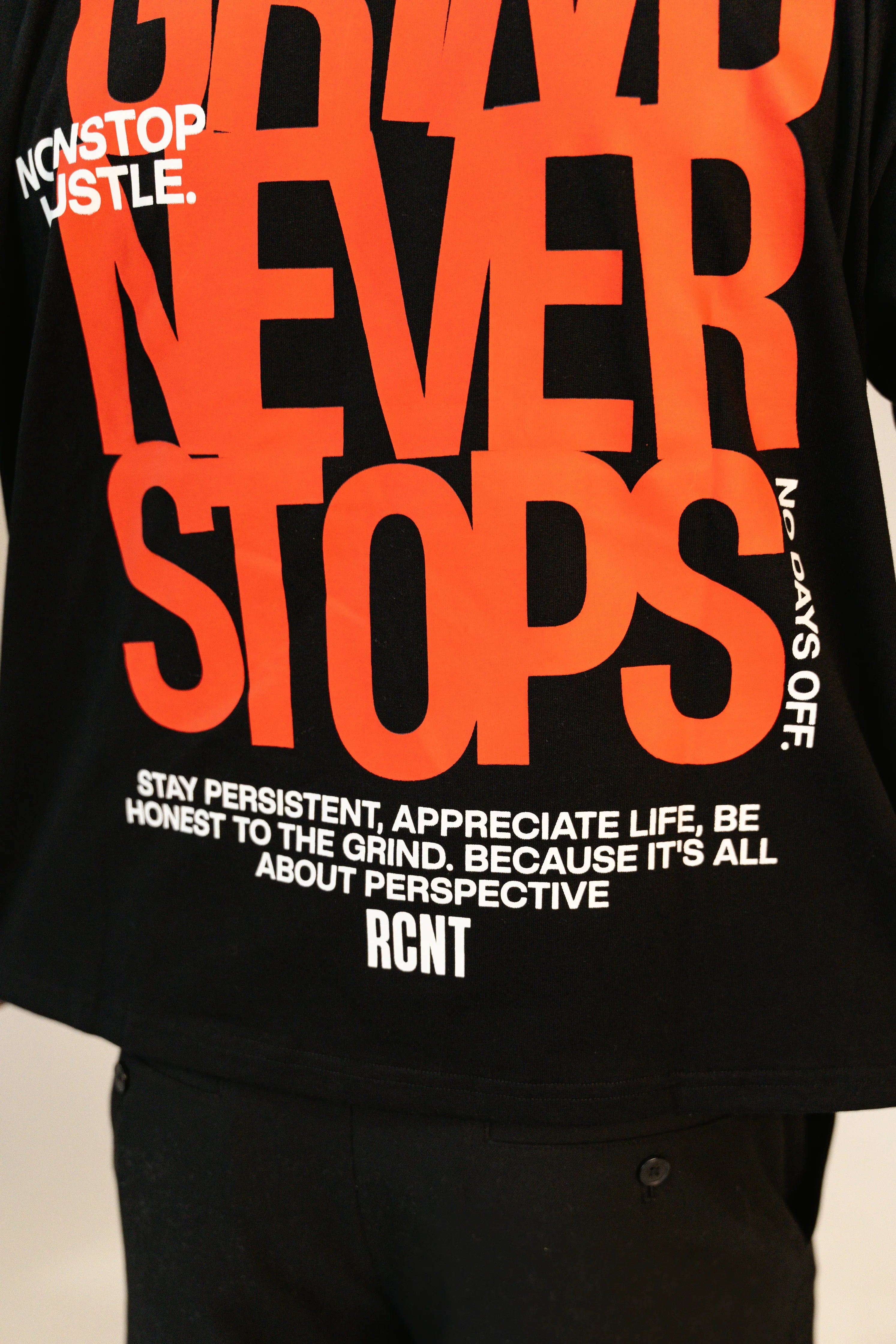 Black T-shirt with bold orange text 'NEVER STOPS' and white motivational phrases about persistence and hustle