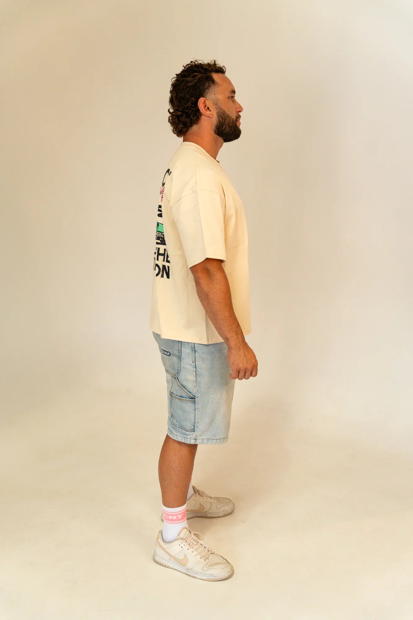 Man in beige graphic T-shirt, light denim shorts, white sneakers, and pink socks standing sideways