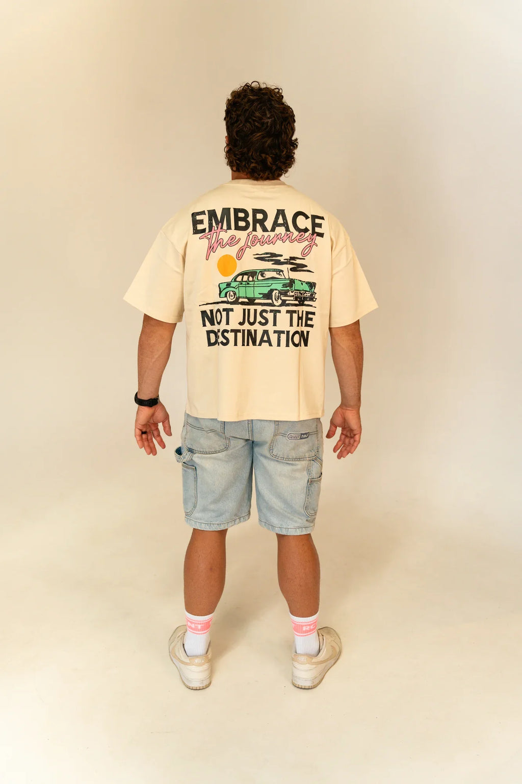 Man wearing beige t-shirt with vintage car and text embracing journey on back, paired with denim shorts