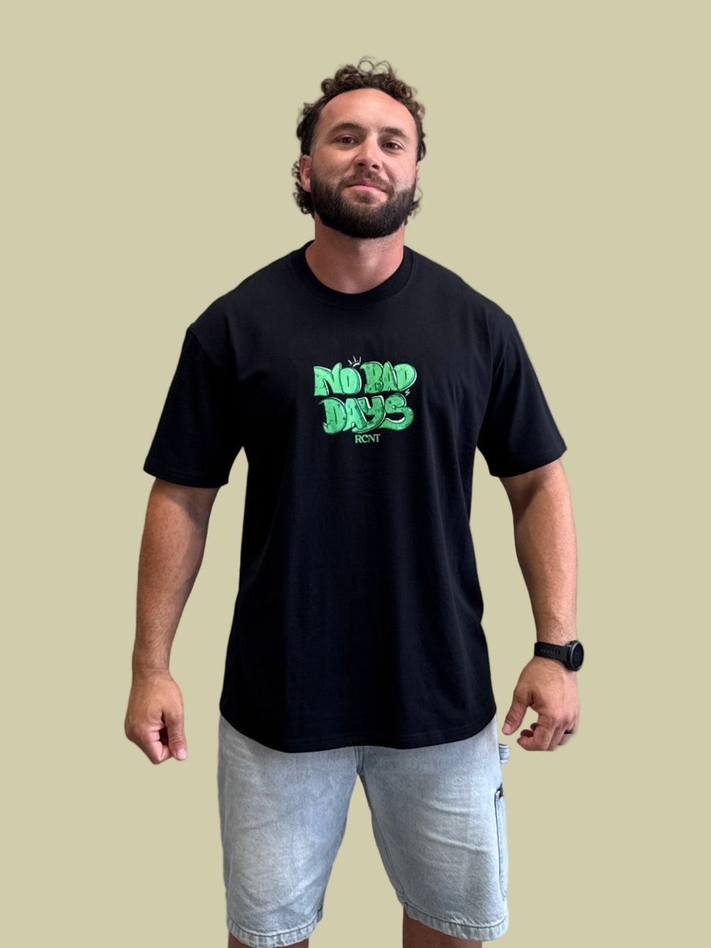 Man wearing black T-shirt with green No Bad Days text and light denim shorts against beige background
