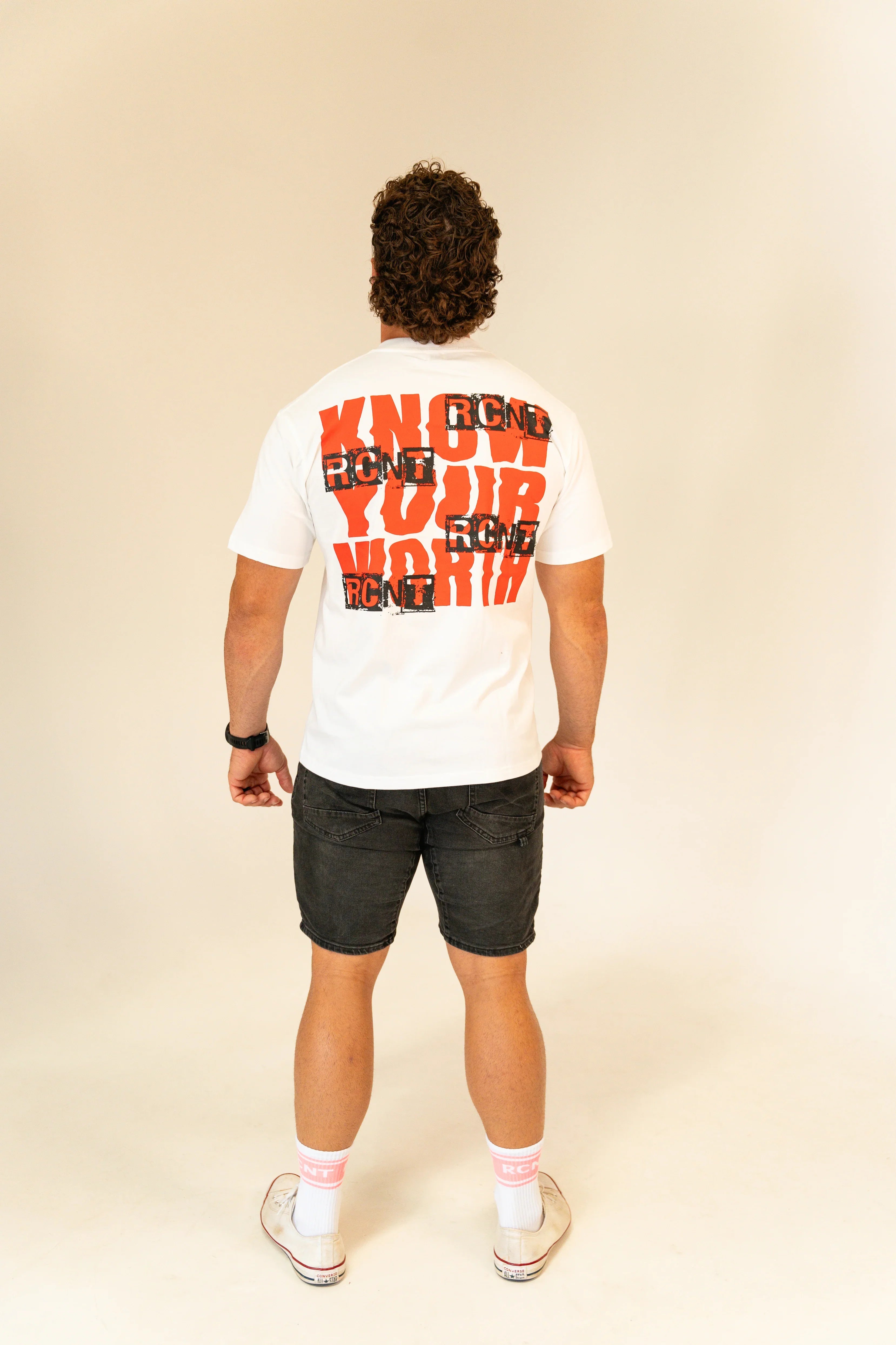 Man wearing white tee with bold red 'Know Your Worth' and black RCNT print, paired with black shorts and white sneakers