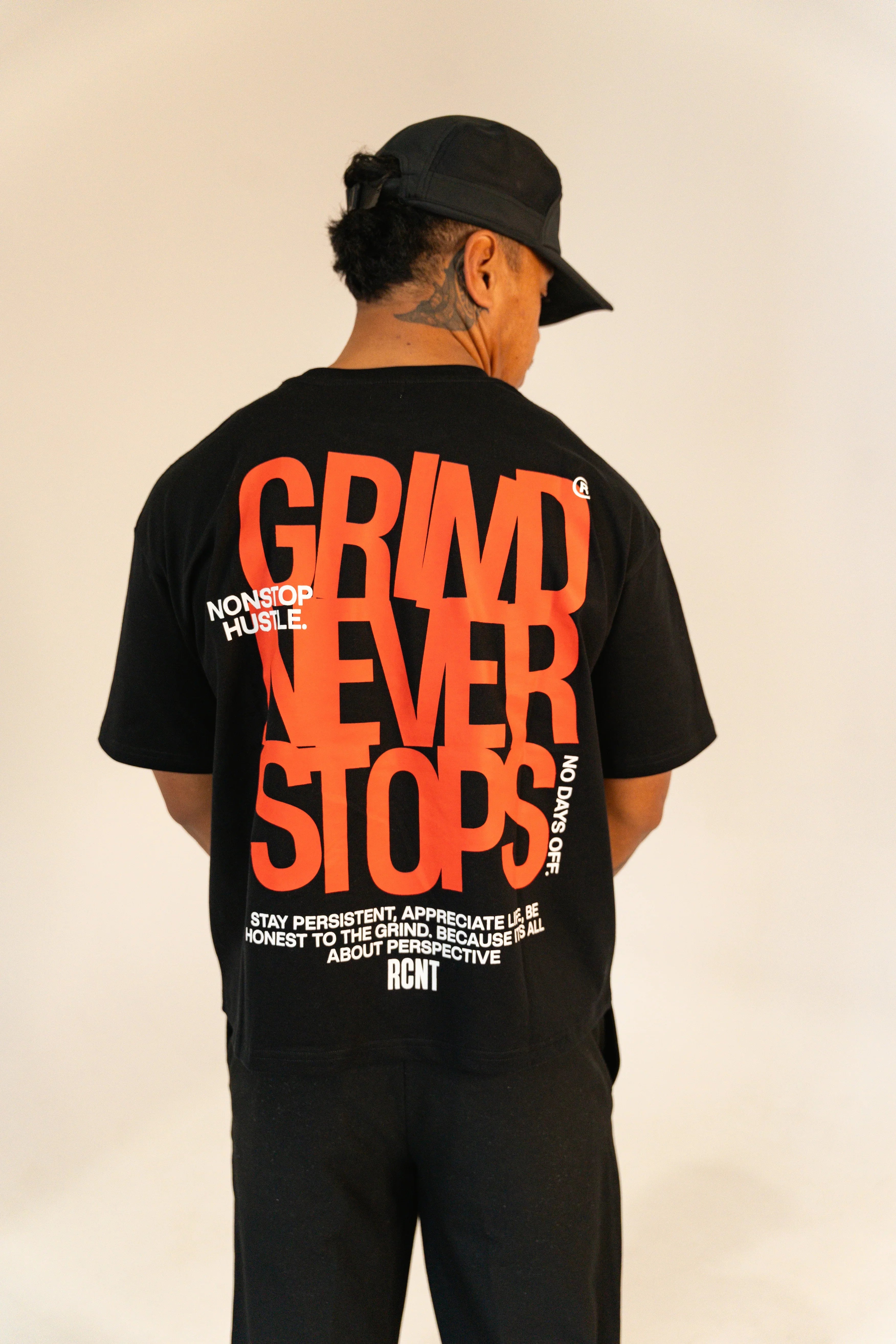 Man wearing black baseball cap and black tee with large red text 'Grind Never Stops' and motivational quotes