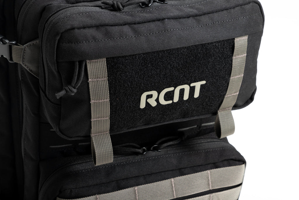 Close-up of black RCNT branded tactical backpack front pocket with gray straps and zipper pulls