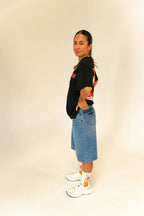 Woman wearing a black graphic t-shirt, knee-length denim shorts, white sneakers, and white-orange socks posing side view