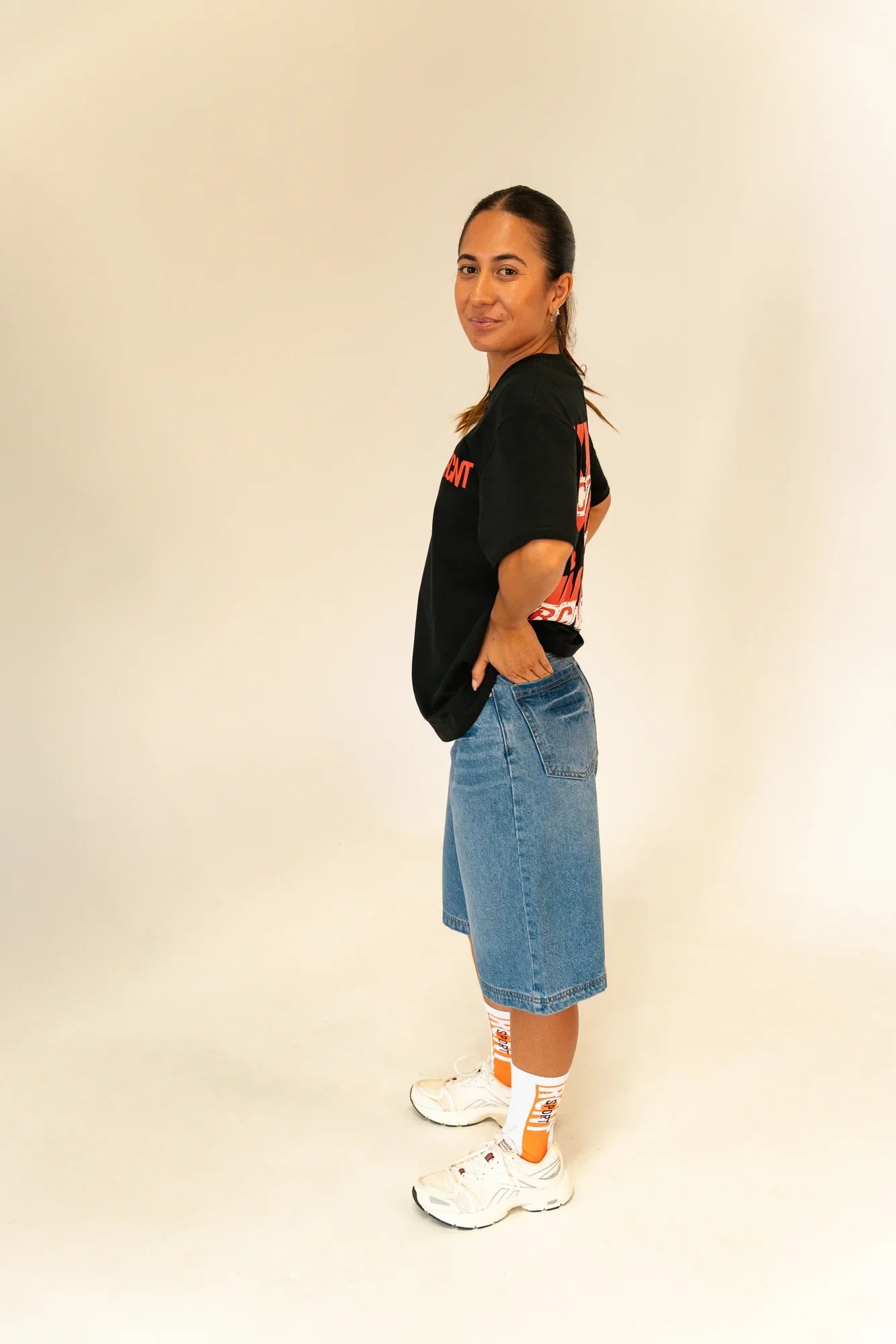 Woman wearing a black graphic t-shirt, knee-length denim shorts, white sneakers, and white-orange socks posing side view