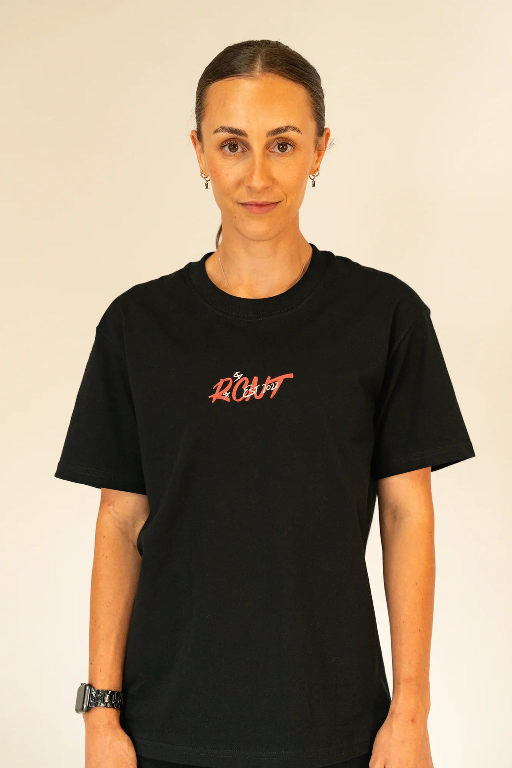 Woman with tied-back hair wearing black t-shirt with red text 'ROOT EST 2022' on plain background