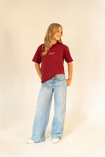 Young woman in maroon graphic t-shirt and wide-leg light blue jeans standing on beige background