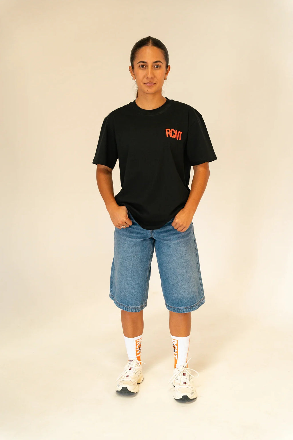 Person wearing black RCMT t-shirt, blue denim shorts, white socks, and white sneakers on plain background