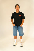 Person wearing black RCMT t-shirt, blue denim shorts, white socks, and white sneakers on plain background