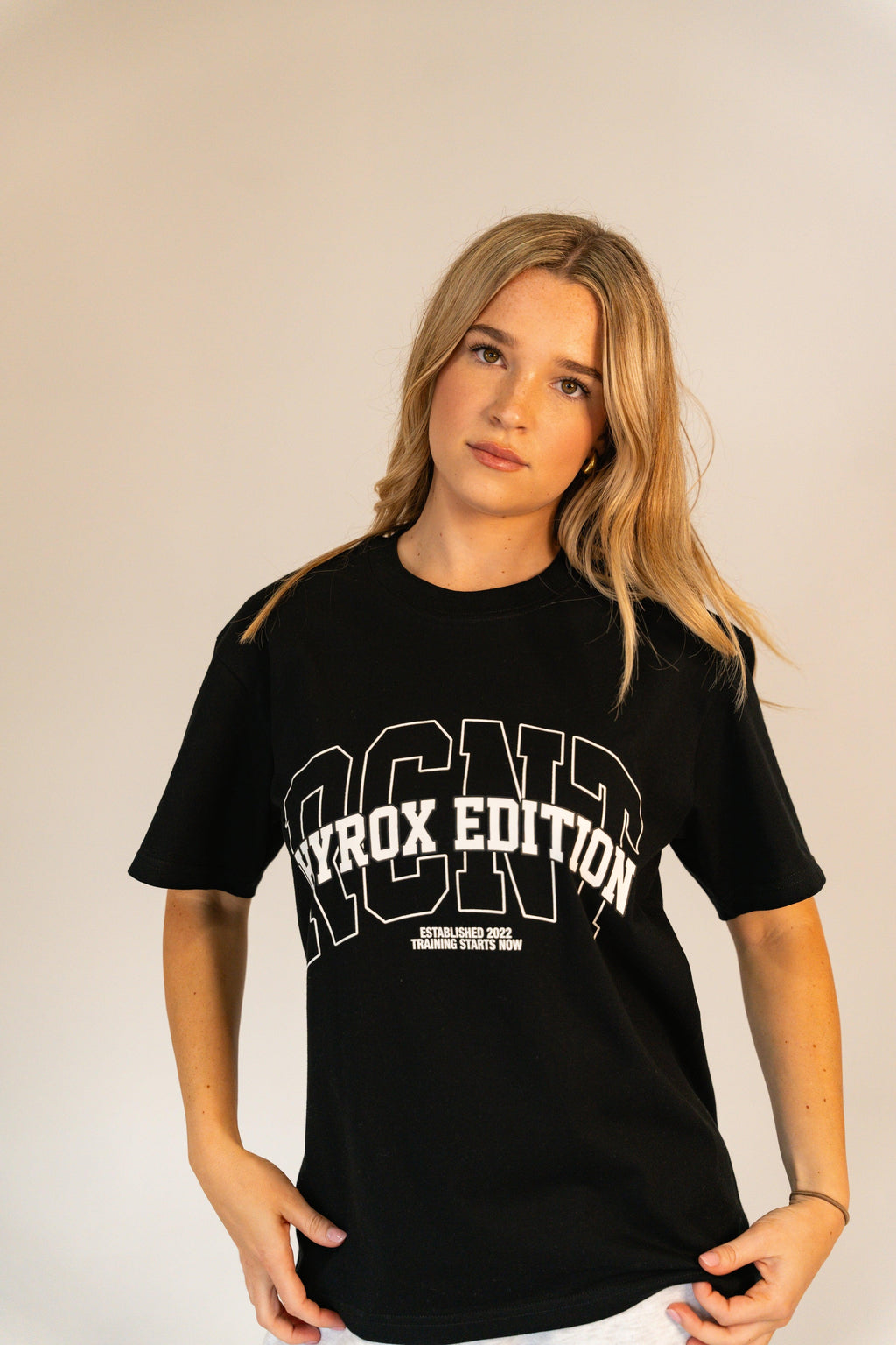 Blonde woman wearing black Hyrox Edition t-shirt posing in front of beige background