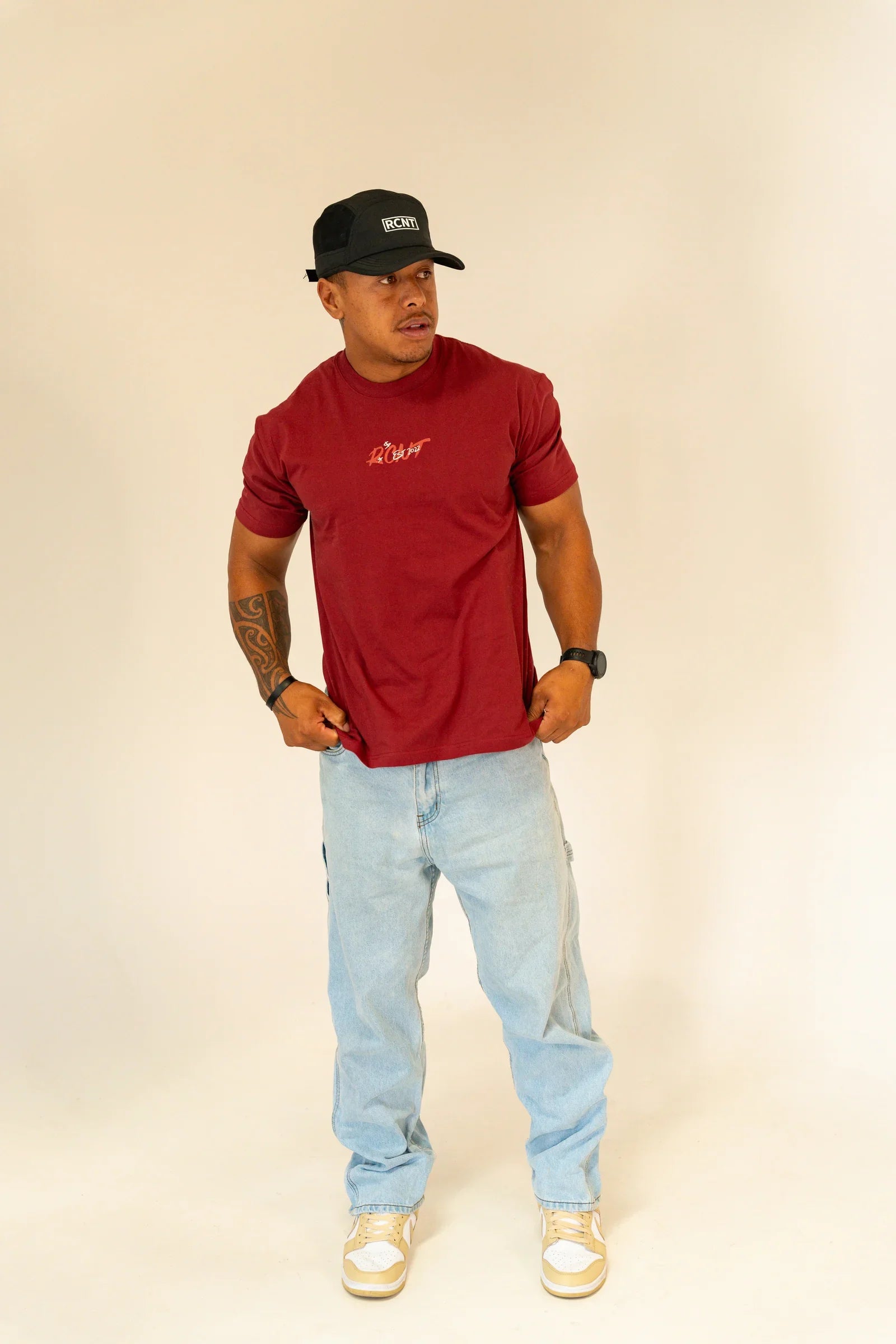 Man in a red RCNT t-shirt, light blue jeans, black RCNT cap, and beige sneakers on white background