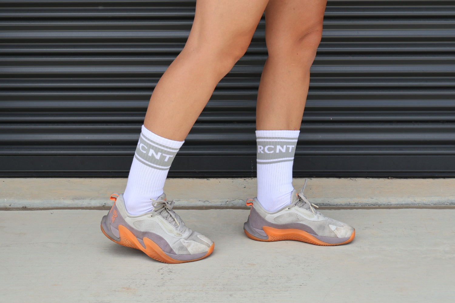 Person wearing white RCNT socks and beige sneakers with orange soles, standing on pavement
