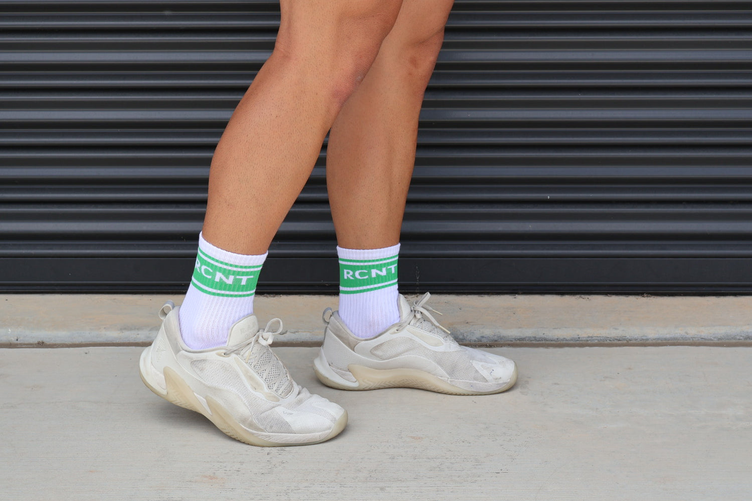 Person wearing white RCNT socks with green stripes and beige sneakers, standing on concrete
