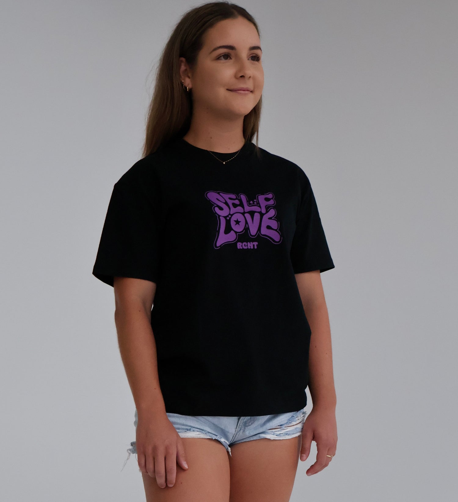 Woman wearing black Self Love graphic t-shirt and denim shorts against plain background