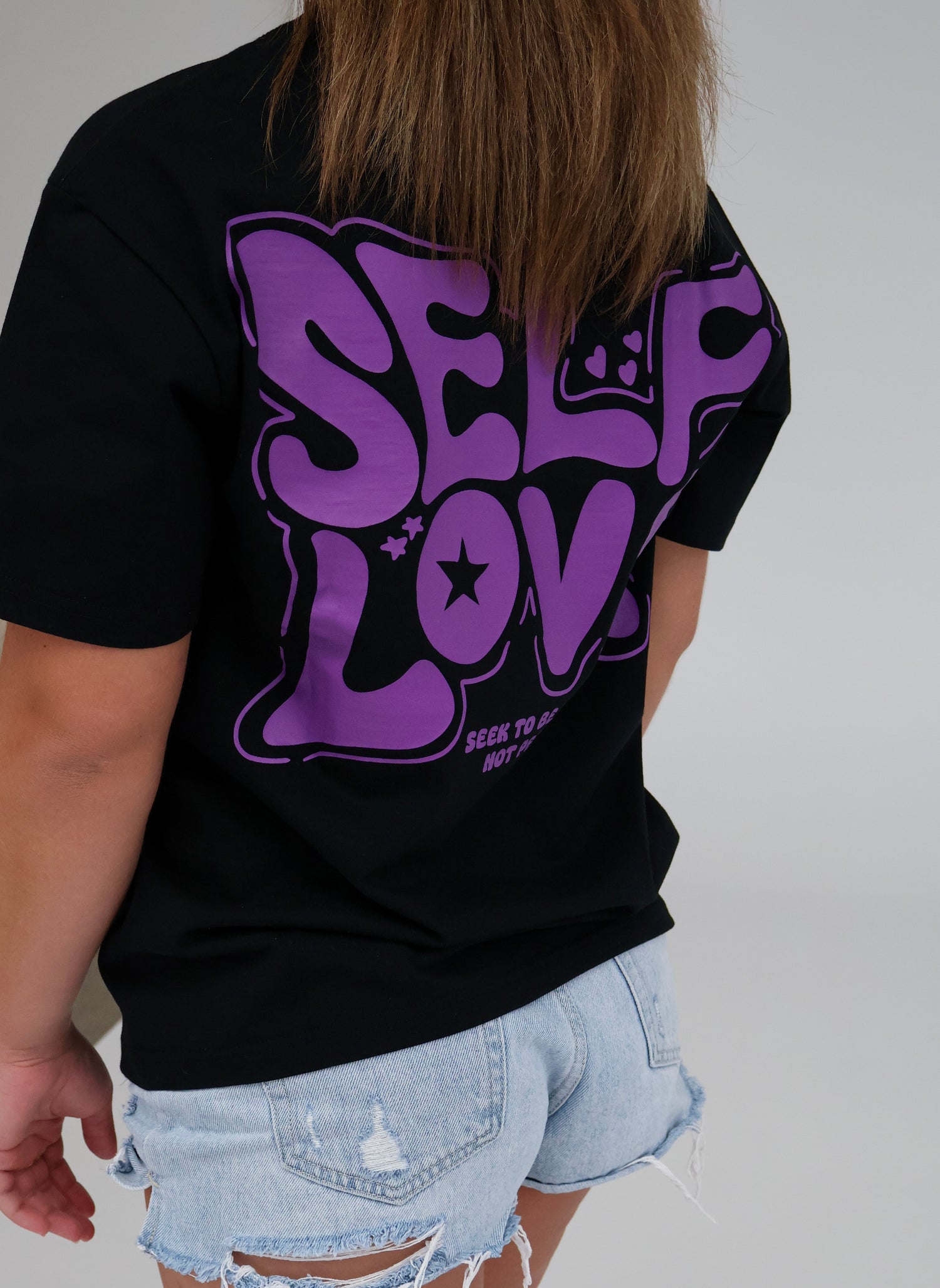 Person wearing black t-shirt with bold purple 'SELF LOVE' graphic and ripped denim shorts