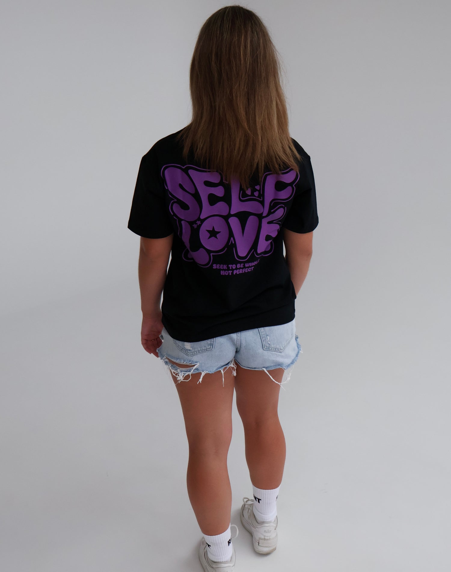 Person wearing black 'Self Love' graphic t-shirt and ripped denim shorts, standing in studio