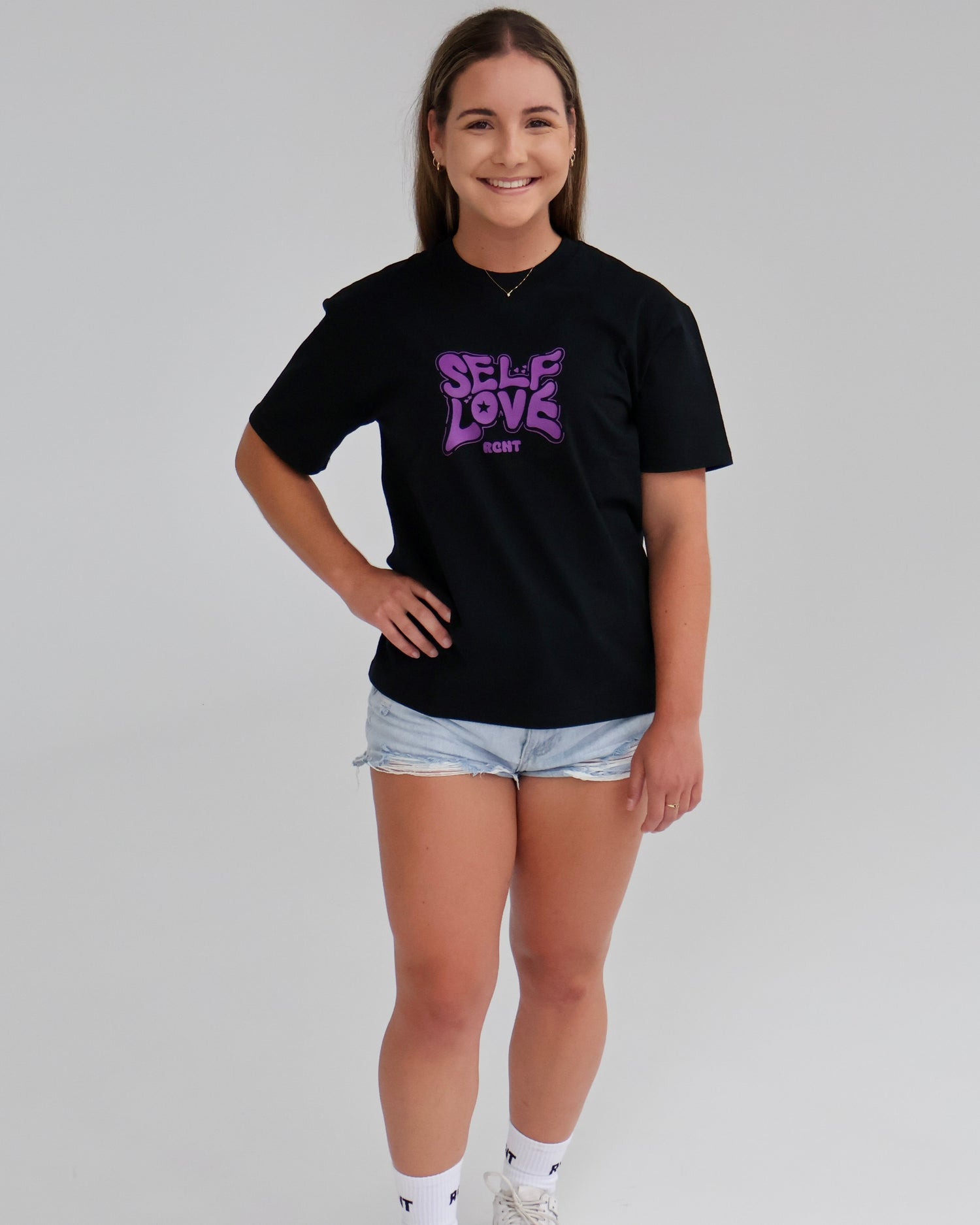 Woman in black Self Love graphic t-shirt, denim shorts, white sneakers against white background