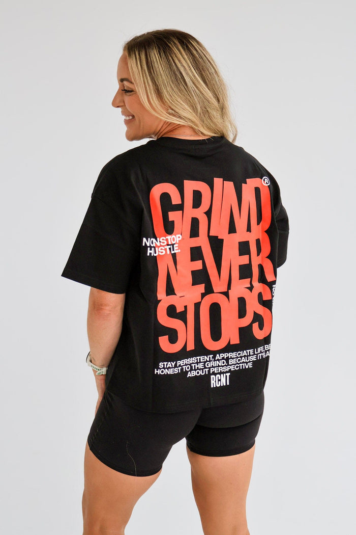 Woman wearing a black t-shirt with 'GRIND NEVER STOPS' text on the back, standing against a plain background.